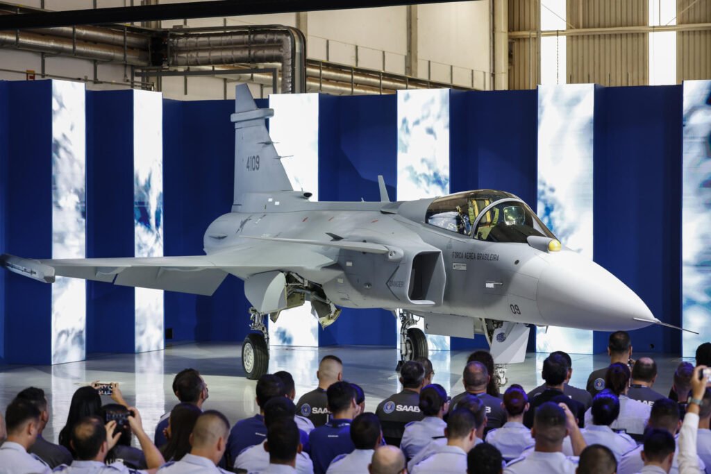 A **Embraer** apresentou o primeiro **caça supersônico** fabricado no Brasil, o **F-39E Gripen**, em uma cerimônia realizada no complexo industrial da empresa em Gavião Peixoto, São Paulo. Este marco é resultado de uma colaboração entre a Embraer, a empresa sueca **Saab** e a **Força Aérea Brasileira (FAB)**. O projeto é parte de um contrato que prevê a produção de 36 caças, dos quais 15 serão montados no Brasil. Este desenvolvimento coloca o Brasil em um grupo seleto de nações capazes de desenvolver e produzir aeronaves de combate de alta complexidade. O que torna o F-39E Gripen especial? Segundo a publicação, o Gripen E é um caça de última geração com 15,2 metros de comprimento e uma envergadura de 8,6 metros. Ele tem um peso máximo de decolagem de 16,5 toneladas e pode atingir velocidades de até 2,4 mil km/h, ou Mach 2, que é aproximadamente duas vezes a velocidade do som. Além disso, o caça possui 10 pontos para fixação de carga externa, permitindo o transporte de até sete mísseis ar-ar Meteor de longo alcance e dois mísseis IRIS-T de curto alcance. Por que a produção nacional é importante? A montagem de 15 das 36 aeronaves em solo brasileiro é crucial para a absorção de conhecimento técnico avançado. Segundo a FAB, essa estratégia faz parte do Programa F-X2 e é fundamental para o desenvolvimento de capacidades locais, desde a produção até o suporte logístico e futuras modernizações. As aeronaves produzidas no Brasil seguem os mesmos padrões de projeto, certificação e qualidade das recebidas da Suécia. Qual é o impacto estratégico do projeto? O ministro da Defesa, José Múcio, destacou que além dos benefícios econômicos e sociais, o projeto do Gripen permite a consolidação do poder dissuasório do Brasil, ampliando a capacidade de garantir a soberania nacional e a segurança regional. Desde fevereiro, o Gripen está a serviço do Alerta de Defesa Aérea, operando a partir da Base Aérea de Anápolis, em Goiás, para proteger o espaço aéreo sobre o Distrito Federal. Como o Gripen E melhora a defesa aérea? A Embraer afirmou que a arquitetura centrada em rede do Gripen E e suas capacidades de fusão de sensores permitem o compartilhamento de informações em toda a formação tática. Isso apoia decisões coordenadas e aprimora a consciência situacional e a resposta a ameaças, tornando o Gripen E uma peça-chave na defesa aérea do Brasil. Quais são os próximos passos do projeto? O contrato assinado em 2014 prevê a entrega de 36 caças, sendo 28 Gripen E monoposto e 8 Gripen F biposto. Até o momento, 11 aeronaves foram entregues, e a expectativa é que a produção continue conforme o cronograma estabelecido. A parceria com a Saab e a produção local são vistas como passos importantes para o fortalecimento da indústria aeronáutica brasileira e para a manutenção da soberania nacional. A presença de autoridades como o presidente Luiz Inácio Lula da Silva e a embaixadora da Suécia no Brasil, Karin Wallensteen, na cerimônia de apresentação, ressalta a importância estratégica e diplomática do projeto. Este desenvolvimento não apenas fortalece a capacidade de defesa do Brasil, mas também representa um avanço significativo na indústria de aviação militar do país.