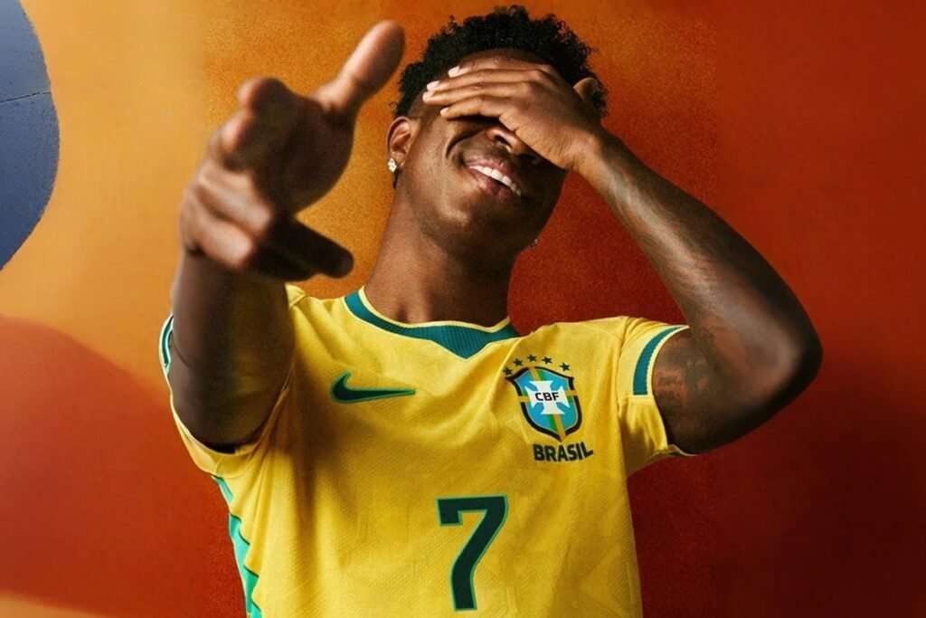 O **uniforme da Seleção Brasileira** para a **Copa do Mundo de 2026** tem sido alvo de diversas polêmicas desde sua concepção. As críticas variam desde a simbologia das cores até a influência estrangeira no design. Segundo a publicação, um dos pontos mais controversos foi a tentativa da Nike e da CBF de introduzir a frase “**Vai, Brasa**” nos itens recém-anunciados. Mas por que essa frase gerou tanto debate? A expressão “**Brasa**” é um apelido para o Brasil, mais comumente utilizado por gerações mais jovens. No entanto, muitos críticos apontam que a palavra não possui um significado claro para a maioria da população. Embora grandes perfis especializados em esportes já tenham usado “Brasa” no passado, o público que realmente entende essa referência é limitado, segundo os críticos. Isso levanta a questão: a escolha de termos modernos e menos conhecidos é eficaz em unir torcedores de diferentes gerações? Outro ponto de discussão é a clássica **camiseta amarela** da Seleção Brasileira, que durante um longo período foi associada ao bolsonarismo e à direita política devido à sua adoção em manifestações. Essa associação impactou o uso da roupa durante a Copa do Mundo de 2026, quando muitas pessoas optaram por versões azuis, verdes ou brancas do uniforme. Anos depois, a camiseta amarela voltou a ter seu significado original, sem grandes associações políticas. No entanto, a cor vermelha ainda carrega um significado ligado à esquerda no imaginário popular. Como essas associações políticas influenciam a percepção e o uso dos uniformes? Em 2025, surgiu a informação de que um dos uniformes oficiais da Seleção para a Copa de 2026 seria na cor da **brasa**, que remete ao nome Brasil. Esse uniforme vermelho teria sido resultado de uma parceria com a **Jordan**, linha de streetwear e basquete da Nike. As reações foram imediatas, com muitos internautas criticando a mudança e especulando sobre uma possível intervenção do governo federal na escolha da cor, especialmente porque a competição ocorre quatro meses antes das eleições presidenciais. Após as críticas, a CBF anunciou que o uniforme para o campeonato não seria vermelho, mas manteria as cores tradicionais: amarelo para o principal e azul para o secundário. Por que a introdução de uma cor tão politicamente carregada gerou tanto alvoroço? Mesmo com a cor vermelha fora dos planos, a Nike manteve a marca **Jordan** como assinatura para o kit reserva da Seleção Brasileira. Isso trouxe influências do streetstyle norte-americano e do basquete nos produtos lançados para a Copa, em parceria com a CBF. A presença de uma personalidade norte-americana, como Michael Jordan, nos uniformes foi vista por muitos como um desrespeito ao Brasil, ao futebol e a Pelé, considerado o maior jogador de futebol da história. Como a presença de ícones de outros esportes e culturas afeta a identidade do futebol brasileiro? Além dos uniformes, a parceria com a Jordan resultou em uma coleção streetwear que inclui camisetas, moletons, shorts e agasalhos. Essa linha também abrange uma coleção infantil, trazendo elementos claros do basquete e da cultura de rua dos Estados Unidos. A campanha focada na estética do futebol de rua foi assinada por Rachel Denti. Como a fusão de diferentes estilos e culturas pode impactar a recepção dos torcedores? Essas questões refletem a complexidade envolvida na criação de um uniforme que não apenas representa um time, mas também carrega significados culturais e políticos profundos.