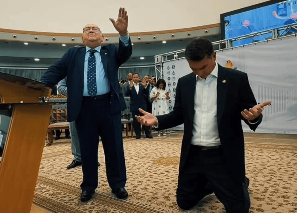 Bastou Flávio Bolsonaro subir ao púlpito para que a cena ganhasse um peso que ia além de uma simples visita religiosa. O que aconteceu ali, em **São Paulo**, e por que esse gesto chamou atenção? Porque o senador e **pré-candidato à Presidência** participou de um encontro de obreiros da **Assembleia de Deus Ministério do Belém** e, diante de lideranças da denominação, recebeu uma oração com menção direta ao seu futuro político. Quando isso ocorreu e em que contexto? A cerimônia foi realizada na **2ª feira, 6 de abril de 2026**, durante um **encontro estadual** da igreja. Não se tratava de uma agenda isolada ou casual. A presença de Flávio marcou o início de uma série de visitas a **igrejas evangélicas no Estado**, em um momento em que a aproximação com esse segmento ganha relevância no cenário eleitoral. Quem conduziu a oração e qual foi o teor da fala? O senador recebeu a oração do **bispo José Wellington Bezerra da Costa**, uma das lideranças presentes no encontro. E foi justamente nesse momento que a dimensão política da cerimônia ficou mais evidente. Durante a oração, o bispo declarou: **“Que o Senhor o leve para ser presidente da nossa nação. Que ele tenha graça e nasça do céu”**. A frase colocou no centro da cena não apenas a presença do parlamentar, mas a associação direta entre fé, liderança religiosa e projeção política. Quem mais participou da reunião? O encontro reuniu cerca de **40 pastores** e diferentes níveis de liderança da denominação. Também esteve presente o **pastor José Wellington Costa Júnior**, filho do bispo. Por que isso importa? Porque ambos são ligados à **Convenção Geral dos Ministros das Igrejas Evangélicas Assembleia de Deus do Brasil**, uma das principais entidades da igreja no país. A participação dessas lideranças reforçou o peso institucional da agenda. A visita terminou ali? Não. A passagem de Flávio pela capital paulista incluiu ainda **encontros reservados com outras lideranças evangélicas**. Quais nomes estavam no radar? Aliados do senador tentavam marcar reuniões com **Estevam Hernandes**, da **Igreja Renascer em Cristo**; **R. R. Soares**, da **Igreja Internacional da Graça de Deus**; e **Valdemiro Santiago**, da **Igreja Mundial do Poder de Deus**. A movimentação, portanto, não se limitou a uma única cerimônia, mas se inseriu em uma agenda mais ampla de aproximação. Por que essa articulação acontece agora? Porque a disputa pelo **eleitorado evangélico** já se intensifica de olho nas **eleições de 2026**. Esse movimento não envolve apenas Flávio Bolsonaro. Na semana anterior, o governador **Ronaldo Caiado (PSD)** lançou uma ofensiva para ampliar sua presença entre os religiosos, com articulação do deputado **Otoni de Paula (MDB)**. O cenário, assim, mostra diferentes atores políticos buscando espaço junto a esse público. E o que, afinal, ficou registrado como ponto mais marcante da agenda em São Paulo? A imagem de **Flávio Bolsonaro** no púlpito da **Assembleia de Deus Ministério do Belém**, cercado por lideranças evangélicas, recebendo a oração do **bispo José Wellington Bezerra da Costa** diante de cerca de **40 pastores**, no início de uma série de visitas a igrejas no Estado. E, no momento mais direto da cerimônia, a fala que resumiu o sentido político daquele encontro: **“Que o Senhor o leve para ser presidente da nossa nação. Que ele tenha graça e nasça do céu.”**