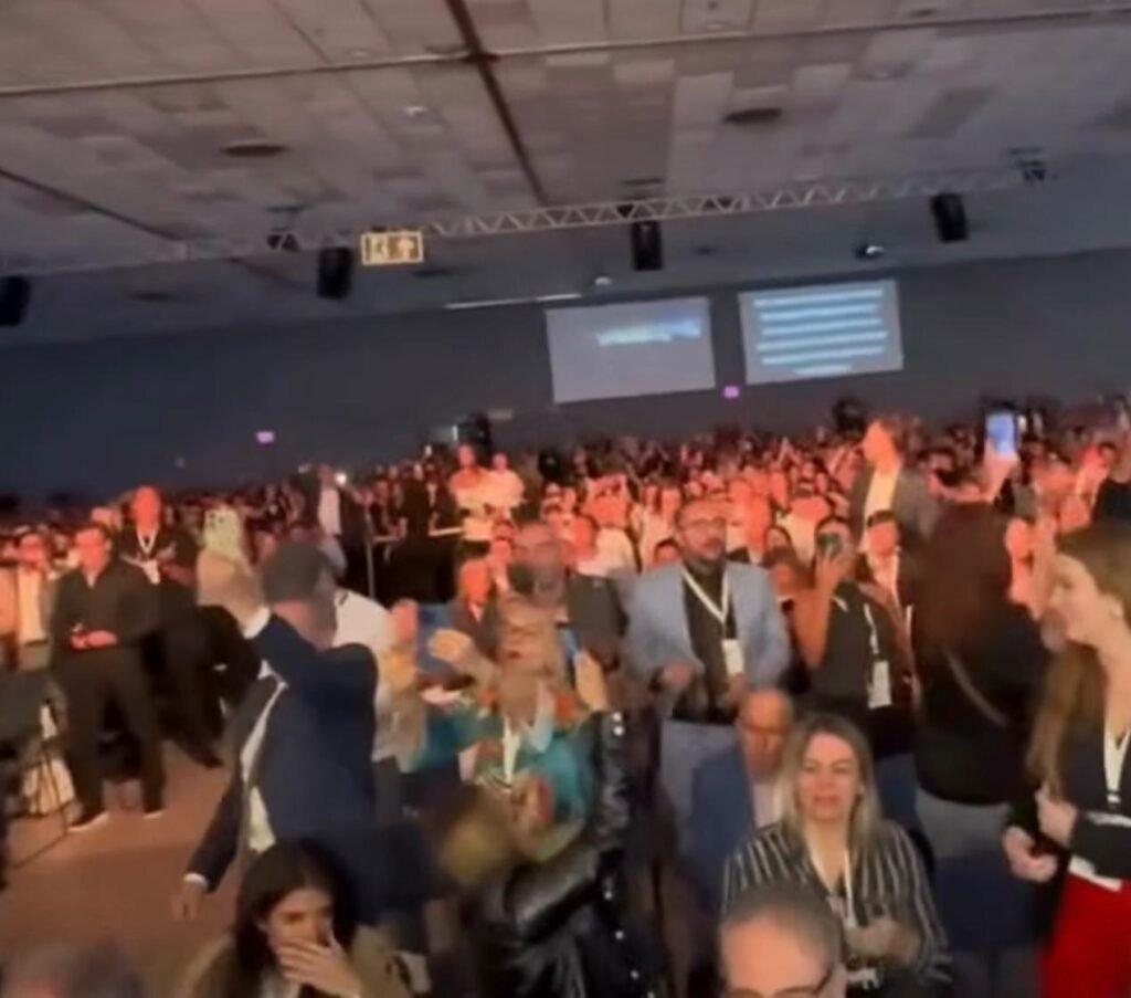 Bastou ele aparecer para o ambiente mudar de tom em segundos. Mas por que uma presença em um evento já conhecido por reunir nomes do debate público provocaria uma reação tão imediata? A resposta começa no peso político de quem chegou ao local, mas não para por aí. O que se viu não foi apenas a participação de um convidado em mais uma agenda pública. Houve uma resposta da plateia, alta, direta e impossível de ignorar. Que resposta foi essa? Em meio à programação, parte do público puxou um coro com forte conteúdo político: “Lula, ladrão, seu lugar é na prisão”. E é justamente esse ponto que transforma uma simples participação em algo maior. Porque quando a reação da plateia rouba a cena, a pergunta deixa de ser apenas quem estava no palco e passa a ser: o que esse momento revela sobre o clima do evento? Antes de responder, há um detalhe que quase ninguém percebe de imediato. Esse tipo de manifestação não acontece no vazio. Ela surge dentro de um ambiente que já carrega expectativas, posicionamentos e tensões do debate nacional. Então, o que parecia ser apenas mais uma presença em uma lista de convidados rapidamente ganhou outro significado. Mas quem era o nome que provocou essa reação? Era o senador Flávio Bolsonaro, do PL. E por que isso importa tanto? Porque sua presença, por si só, já atrai atenção em qualquer espaço onde política e opinião pública se cruzam. Só que o episódio não se resume ao senador. O que acontece ao redor dele é que passa a concentrar o olhar de quem acompanha. E onde tudo isso aconteceu? Foi durante o Fórum da Liberdade, realizado em Porto Alegre, nesta sexta-feira. Flávio integrou a lista de mais de 40 palestrantes convidados do evento. A princípio, isso poderia soar como apenas mais um nome entre muitos. Só que é aqui que muita gente se surpreende: em certos momentos, não importa quantos palestrantes estejam na programação. Basta uma presença específica para alterar completamente a temperatura do ambiente. Então o foco estava no discurso dele? Não exatamente. O que marcou sua passagem foi a manifestação política vinda de parte da plateia. E isso levanta outra questão: por que esse coro ganhou tanta relevância? Porque ele condensou, em poucos segundos, o tom político que atravessou aquele instante. Não foi uma reação discreta, nem algo periférico. Foi um sinal claro de como o público presente escolheu se expressar. Mas isso aconteceu isoladamente, como um episódio solto dentro da programação? Pelo que foi descrito, ocorreu em meio às atividades do fórum, que reúne lideranças e nomes do debate público para discutir temas econômicos e políticos. E esse contexto muda tudo. Quando uma manifestação assim surge dentro de um evento voltado justamente à discussão pública, ela deixa de ser apenas ruído e passa a funcionar como retrato de um ambiente mais amplo. Só que ainda fica uma dúvida importante: o que esse momento realmente expõe? Expõe que, mesmo em espaços organizados para debates formais, a reação da plateia pode se tornar o fato central. E mais do que isso, mostra como certos nomes políticos carregam consigo uma capacidade imediata de mobilizar sentimentos, palavras de ordem e disputas simbólicas. Mas há outro ponto que chama atenção. O episódio não foi apresentado como um confronto direto no palco, e sim como uma manifestação da plateia durante a presença do senador. Isso faz diferença? Faz, porque desloca o centro da cena. Em vez de um embate verbal tradicional, o que se destacou foi a resposta espontânea de parte do público, dando ao momento um peso político próprio. No fim, o principal não foi apenas que Flávio Bolsonaro esteve no Fórum da Liberdade, em Porto Alegre. O que realmente marcou sua participação foi o coro entoado por parte da plateia contra o presidente Lula, transformando sua presença em um episódio político dentro da programação do evento. E o que isso ainda pode revelar sobre o clima desses encontros públicos talvez comece justamente onde a notícia para.