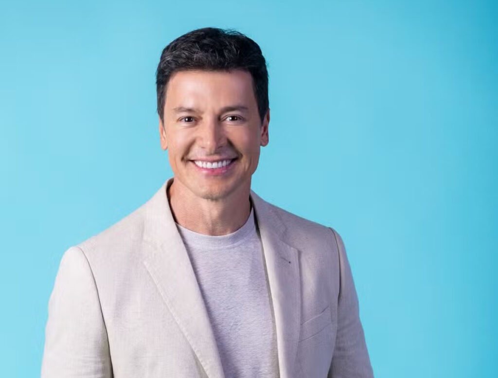 Não será apenas mais um reality: **Rodrigo Faro** volta à **Globo** para comandar um jogo em que a disputa passa por **manipulação**, **traições** e **tensão psicológica**. Mas o que, de fato, está por trás de **“Herança em Jogo”**? A resposta começa pelo próprio formato, pensado para colocar competidores diante de escolhas em que **inteligência**, **poder de persuasão** e **pensamento estratégico** valem tanto quanto a capacidade de sustentar alianças frágeis. E como essa dinâmica funciona? A proposta mistura **ficção e realidade** em um ambiente de pressão, onde os participantes precisam decidir até onde estão dispostos a **mentir** ou **trair** para seguir na disputa. Não se trata apenas de resistir ao confinamento ou vencer provas isoladas. O centro da experiência está no jogo psicológico, nas relações instáveis e na forma como cada decisão pode alterar o rumo da competição. Qual será o papel de Rodrigo Faro nesse cenário? Ele não aparecerá apenas como um apresentador convencional. Em **“Herança em Jogo”**, Faro assumirá a função de **“testamenteiro”**, descrita como a de condutor oficial da dinâmica. E o que isso significa na prática? Significa que ele será o responsável por garantir o cumprimento do que foi estabelecido dentro do jogo, ocupando uma posição diretamente ligada às regras e ao andamento da disputa. O próprio Faro resumiu essa função de forma clara. “Como **testamenteiro**, meu papel é garantir que cada linha do que foi documentado seja honrada, mas confesso: o que mais me intriga é ver até onde os participantes serão capazes de ir para conquistar esse jogo”, afirmou ele em nota. Essa declaração ajuda a entender o tom da atração: menos leveza, mais pressão; menos condução neutra, mais presença dentro da lógica do programa. Por que essa estreia chama tanta atenção? Porque ela marca o retorno de **Rodrigo Faro à Globo com um vínculo fixo após 18 anos**. A confirmação também acontece depois do fim do **“Hora do Faro”**, exibido entre **2014 e 2024**. Isso transforma o novo projeto em um movimento importante na trajetória do apresentador, agora associado a uma plataforma que vem reforçando sua aposta em realities. Mas **“Herança em Jogo”** nasceu no Brasil? Não. A produção brasileira é uma adaptação de **“The Inheritance”**, formato criado pela produtora britânica **Studio Lambert**, conhecida por desenvolver sucessos globais do gênero. E há outro detalhe relevante: esta será a **primeira versão internacional** do programa, o que coloca a edição brasileira em uma posição estratégica dentro da expansão da franquia. Como era o formato original? Na versão britânica, o posto equivalente ao de Faro era ocupado pelo juiz **Rob Rinder**, o que dava à disputa um tom mais **verídico** e **legalista**. O elenco também contava com a atriz **Elizabeth Hurley**, conhecida por seu relacionamento com **Hugh Grant** nos anos 1990. Esses elementos ajudavam a compor a identidade da edição inglesa, que agora servirá de base para a adaptação nacional. E o original foi um sucesso? Não. Apesar da expectativa em torno do lançamento pelo **Channel 4**, no ano passado, a produção enfrentou **custos elevados** e registrou **baixa audiência no Reino Unido**. O resultado foi o **cancelamento precoce após a primeira temporada**. Esse histórico naturalmente levanta dúvidas sobre o desempenho da versão brasileira. Então por que a Globo aposta no projeto? Porque a avaliação interna é de **otimismo** diante do potencial de engajamento do público brasileiro com **formatos de confinamento**. A produção nacional ficará a cargo da **Formata**, com **gravações programadas para começar em maio** e **estreia prevista no catálogo do Globoplay ainda neste ano**. A plataforma vê os realities como parte central de sua estratégia. Essa confiança foi resumida por **Tatiana Costa**, diretora de Canais de Entretenimento e Conteúdo de Produtos Digitais da Globo: “Os **realities** são um dos principais pilares do **Globoplay**, e acreditamos fortemente no potencial deste formato para criar conexão e engajar o público brasileiro”. A percepção é compartilhada também por quem distribui o formato internacionalmente. O que diz a distribuidora? A **All3Media International** afirma confiar na experiência da emissora brasileira para mudar o histórico da franquia. “O histórico da **Globo** na entrega de realities imperdíveis faz dela a parceira ideal para levar este formato ousado e distinto às telas brasileiras”, justificou **Yari Torres**, vice-presidente da empresa para a América Latina. E, afinal, como será **“Herança em Jogo”** na **Globoplay**? Será um reality de **confinamento** centrado em **manipulação**, **traições** e **tensão psicológica**, adaptação brasileira de **“The Inheritance”**, com **Rodrigo Faro** no papel de **testamenteiro**, produção da **Formata**, gravações a partir de **maio** e estreia no **Globoplay ainda neste ano**.