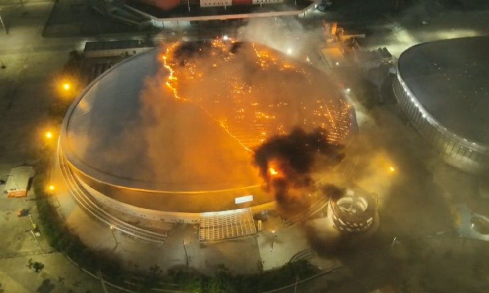 O fogo começou antes do amanhecer e atingiu um dos espaços mais simbólicos do legado olímpico do Rio. Onde isso aconteceu? No **telhado do Velódromo do Parque Olímpico**, na **Barra Olímpica**, na **Zona Sudoeste do Rio de Janeiro**, durante a madrugada desta **quarta-feira (8)**. Quando as equipes foram chamadas? O **Corpo de Bombeiros Militar do Estado do Rio de Janeiro** informou que foi acionado às **4h17** para combater as chamas. O que se sabia naquele momento? Que o incêndio estava concentrado na parte superior da estrutura e exigia uma operação ampla para controle da situação. Havia vítimas? De acordo com a corporação, **não há vítimas**. E o interior do velódromo foi atingido? Segundo os bombeiros, a **estrutura interna não foi danificada**. Isso significa que o fogo não se espalhou para outras áreas do complexo? Até o fim da manhã, a informação oficial era de que **não havia risco de propagação**. E o que aconteceu com o restante do Parque Olímpico? Um dos pontos de maior atenção era o **Museu Olímpico**, que faz parte do complexo. Ele foi afetado? A corporação informou que o museu foi **preservado**. Mais tarde, em entrevista à mídia local, o prefeito do Rio, **Eduardo Cavaliere**, reforçou que os danos foram limitados. O que exatamente ele disse? Que o museu está **praticamente preservado** e que o **Velódromo** e o **Museu Olímpico** **quase não foram impactados**. Então nada foi atingido? Houve, sim, um impacto pontual. Segundo Cavaliere, **apenas uma sala imersiva foi impactada**. Por que os danos foram contidos? O prefeito destacou a atuação da equipe local e afirmou que a **brigada do local** foi fundamental para, junto aos bombeiros, **minimizar os efeitos do incêndio**. Quantas equipes foram mobilizadas para a ocorrência? A operação reuniu cerca de **80 bombeiros**, de **seis unidades**, com apoio de **mais de 20 viaturas** e **equipes especializadas**. Até quando o trabalho seguia em andamento? Às **9h17** desta quarta-feira, os bombeiros ainda atuavam no combate às chamas e no controle da situação. Havia previsão para o fim da ocorrência? **Não havia previsão**. As causas do incêndio já foram identificadas? Ainda não. Segundo as informações divulgadas, as **causas são desconhecidas** e serão **apuradas após o término do combate ao fogo**. Houve interdição no trânsito da região? **Não há bloqueios nas vias do entorno**, de acordo com a corporação. Por que o velódromo chama tanta atenção? Porque se trata de uma estrutura construída para os **Jogos Olímpicos de 2016**, destinada às competições de **ciclismo de pista**. Ele sempre esteve ali? Não. O atual velódromo **substituiu o antigo velódromo do Pan de 2007**, que foi demolido por **não atender às exigências internacionais**. Qual é o porte dessa arena? O espaço tem capacidade para cerca de **5 mil espectadores** e conta com **pista de madeira voltada ao alto rendimento**. Quanto custou a obra? Cerca de **R$ 140 milhões**. A construção foi tranquila? Não. As obras enfrentaram **preocupação constante por causa de repetidos atrasos**, inclusive na entrega. Quando o velódromo foi inaugurado? Em **26 de junho de 2016**. Houve atraso nessa entrega? Sim, a inauguração ocorreu com **seis meses de atraso**. E ele chegou a receber evento-teste antes da Olimpíada? Não. Foi o **único equipamento dos Jogos Olímpicos de 2016** que **não passou por um evento-teste**. O que permanece confirmado até agora? Que o incêndio atingiu o **telhado do Velódromo no Parque Olímpico**, que o chamado aos bombeiros ocorreu às **4h17**, que **não há vítimas**, que a **estrutura interna não foi danificada**, que o **Museu Olímpico foi preservado**, que **não há risco de propagação**, que cerca de **80 bombeiros**, de **seis unidades**, atuam com **mais de 20 viaturas**, que **não há bloqueios nas vias do entorno**, que as **causas ainda serão apuradas**, e que, segundo **Eduardo Cavaliere**, os danos foram **limitados**, com impacto em **apenas uma sala imersiva**.