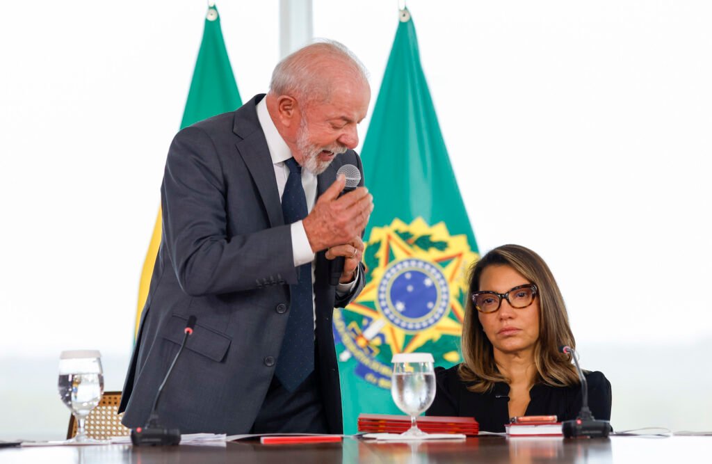 Uma palavra bastou para mudar o peso inteiro de uma cena que, à primeira vista, parecia apenas protocolar. Mas que palavra foi essa? E por que ela chamou tanta atenção justamente em um evento sobre **proteção à mulher**? A resposta começa em um momento público, diante de autoridades, quando o presidente **Lula** mencionava uma operação da **Polícia Federal** que havia resultado na prisão de cerca de **5.000 pessoas** por **violência de gênero**. Só que eram mesmo “pessoas”? É aí que o episódio ganha outra dimensão. Antes que a frase seguisse adiante como mais uma estatística de discurso oficial, houve uma interrupção curta, direta e impossível de ignorar. A correção veio de **Janja**, que trocou o termo genérico por uma definição mais precisa: **“5.000 homens”**. Por que essa troca importa tanto, se o número continuava o mesmo? Porque, em temas como **violência contra a mulher**, a escolha das palavras não é detalhe secundário. Quando se diz “pessoas”, a frase parece diluir o perfil de quem comete o crime. Quando se diz “homens”, o foco muda. A responsabilidade deixa de ficar abstrata. E é justamente aqui que muita gente se surpreende: não foi uma correção sobre quantidade, mas sobre **sentido**. E o que aconteceu depois? O próprio presidente retomou a fala já com a alteração incorporada: **“Em um único dia, nós prendemos quase 5.000 homens por violência contra a mulher”**. A cena, então, deixou de ser apenas uma interrupção e passou a simbolizar algo maior. Mas maior em que sentido? A dúvida cresce porque esse não foi um episódio isolado em torno dos discursos de Lula. Nos bastidores e também em público, o presidente tem recebido **intervenções pontuais** e **recomendações prévias** de auxiliares do **Planalto**. Isso significa controle maior sobre a comunicação? Em parte, sim. Mas há um ponto que quase passa despercebido: quanto mais o presidente fala, mais ele também fica exposto. E por que essa exposição aumentou? Desde que **Sidônio Palmeira** assumiu a **Secom**, a estratégia adotada foi fazer com que Lula falasse mais e concedesse mais entrevistas. A intenção era ampliar presença, reforçar mensagem, ocupar espaço. Só que o efeito colateral veio junto. Com mais falas públicas, cresceram também as chances de **gafes**, **lapsos** e declarações controversas. Isso já tinha acontecido antes? Sim, e esse é o detalhe que reacende a curiosidade no meio da história. Em **Salvador**, por exemplo, Sidônio pediu ao presidente que comentasse sobre o **Pix**. O esperado era uma fala objetiva sobre o tema. O que veio, porém, tomou outro rumo e acabou em críticas ao presidente dos **Estados Unidos**, **Donald Trump**. O que parecia orientação de comunicação virou desvio de foco. Então a correção de Janja foi só um ajuste pontual? Talvez tenha sido mais do que isso. Um levantamento do **Poder360** apontou que, em **3 anos de governo**, Lula deu ao menos **157 declarações** com distrações ou incorreções. Esse número, por si só, já levanta outra pergunta: quando uma correção acontece em público, ela constrange ou protege? A resposta depende do olhar. Para alguns, expõe fragilidade. Para outros, evita que o erro se consolide. E por que esse episódio específico repercute tanto? Porque ele ocorreu durante a **sanção de leis de proteção à mulher**, um contexto em que cada palavra ganha peso político e simbólico. Não era apenas uma cerimônia. Era um momento em que o discurso precisava estar alinhado ao tema central. E é aqui que tudo muda: a correção não mexeu só na frase, mexeu na forma como o problema foi enquadrado diante do público. No fim, o que ficou não foi apenas a imagem de **Janja corrigindo Lula**, mas a força de uma intervenção que reposicionou a mensagem no instante exato em que ela poderia se perder. E o ponto principal aparece justamente aí, quase no fim: ao trocar **“pessoas”** por **“homens”**, Janja recolocou no centro da fala quem, segundo o relato do evento, havia sido preso por **violência contra a mulher**. Só que a cena não se encerra nessa correção. Ela também deixa no ar uma pergunta que continua aberta: em um governo que decidiu expor mais a voz do presidente, quantas vezes uma única palavra ainda será capaz de redefinir toda a narrativa?