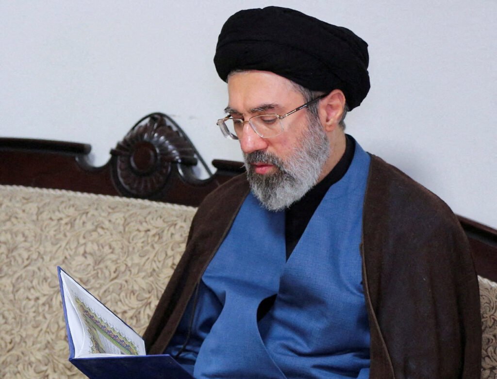 O homem que assumiu o posto mais poderoso do Irã estaria **inconsciente**, longe dos holofotes e sem condições de governar. Quem é esse nome no centro da crise? Trata-se de **Mojtaba Khamenei**, apontado como o novo **líder supremo do Irã**, segundo informações publicadas pelo jornal **The Times**. Mas o que exatamente foi relatado? De acordo com o jornal, Mojtaba está **incapacitado** e recebendo **tratamento médico** na cidade sagrada de **Qom**, com base em uma avaliação da inteligência dos **Estados Unidos** e de **Israel** à qual o veículo teve acesso. Por que isso chama tanta atenção? Porque, segundo o memorando diplomático citado, ele estaria em estado **grave** e “incapaz de participar de qualquer tomada de decisão do regime”. Onde ele estaria sendo tratado? O local indicado é **Qom**, cidade central situada a cerca de **140 quilômetros ao sul de Teerã** e considerada um lugar sagrado no **islamismo xiita**. E por que essa informação pesa tanto? Porque o líder supremo, no sistema iraniano, ocupa a mais alta autoridade **política** e **religiosa** do país. Se ele não pode atuar, a dúvida sobre quem realmente conduz o poder se torna inevitável. Como essa situação começou? O próprio Irã já havia confirmado anteriormente que Mojtaba foi **ferido** no mesmo **ataque aéreo** que matou seu pai, sua mãe, sua esposa **Zahra Haddad-Adel** e um de seus filhos, no primeiro dia da guerra entre **EUA**, **Israel** e **Irã**. O que aconteceu depois disso? Desde então, o sucessor de Khamenei **não foi visto em público**. Houve alguma manifestação atribuída a ele? Segundo o **The Times**, apenas **duas declarações** atribuídas ao líder foram lidas na **televisão estatal iraniana**. Além disso, circulou um **vídeo produzido por inteligência artificial** em que Mojtaba entra em uma sala de guerra e analisa a usina nuclear de **Dimona**, em Israel. Isso esclarece sua condição? Não. Ao contrário, a ausência pública e o uso desse material ampliaram as dúvidas. Quem estaria no controle, então? O jornal afirma que a suposta incapacidade de Mojtaba intensifica as especulações de que a **Guarda Revolucionária Islâmica** mantém o comando do país e que ele funcionaria como um **fantoche**. Há algum sinal político nessa direção? Sim. **Donald Trump** afirmou que está negociando com **funcionários iranianos**, e não com o líder supremo. O cenário interno também mudou? Segundo a reportagem, o regime iraniano convocou **soldados aposentados**, enquanto a força paramilitar **Basij**, ligada à Guarda Revolucionária Islâmica, começou a aceitar combatentes com pelo menos **12 anos de idade**. Houve resposta oficial às informações? O **The Times** procurou a **Agência de Segurança Nacional dos EUA**, a **NSA**, e também a representação do Irã em Washington, mas não obteve retorno. E por que o momento é ainda mais sensível? Porque na quarta-feira, **8 de abril**, completam-se **40 dias** desde a morte de **Ali Khamenei**, ocorrida em **28 de fevereiro**, data que marcaria o fim do período tradicional de luto no islamismo xiita. O que mais foi informado sobre isso? O memorando citado pelo jornal também afirma que o corpo de **Ali Khamenei** estaria sendo preparado para um enterro em **Qom**. Por que isso também levanta questionamentos? Porque a tradição xiita prevê um **sepultamento rápido**, mas o funeral ainda não ocorreu. Qual foi a justificativa do governo iraniano? Segundo a versão oficial, a cerimônia foi adiada pela “**expectativa de uma participação sem precedentes**”. No fim, o que está confirmado no relato publicado? Que **Mojtaba Khamenei**, novo **líder supremo do Irã**, está em **Qom**, sob **tratamento médico**, em estado **grave**, **inconsciente** e **incapaz de participar de qualquer tomada de decisão do regime**, segundo informações divulgadas pelo **The Times** com base em avaliação da inteligência dos **Estados Unidos** e de **Israel**.