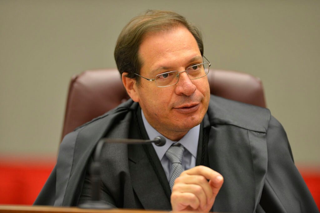 Uma votação unânime, em um tribunal cercado por crises, parece sinal de força ou de urgência? A resposta imediata aponta para força institucional. Quando quase todos caminham na mesma direção, a imagem transmitida é a de estabilidade. Mas por que essa unanimidade chama tanto a atenção agora? Porque ela não aconteceu em um momento de calmaria, e sim quando a Corte tenta atravessar um dos períodos mais delicados de sua imagem pública. Que momento é esse? O tribunal lida com um inquérito sobre venda de sentenças envolvendo advogados e ex-assessores de ministros. E, no mesmo dia da eleição, ainda precisou tratar de denúncias de assédio sexual contra um de seus integrantes. Então a escolha foi apenas administrativa? Não exatamente. Ela também carrega um peso simbólico, e é aí que muita gente começa a olhar com mais atenção. Se a eleição foi simbólica, o que ela quis mostrar? Segundo o atual presidente da Corte, a decisão unânime buscou demonstrar uma forte união da instituição diante das crises. Mas há um ponto que quase passa despercebido: quando a necessidade de mostrar união é declarada de forma tão explícita, a pergunta deixa de ser apenas quem venceu e passa a ser por que essa mensagem precisava ser enviada com tanta clareza. E quem foi escolhido para carregar esse recado? O ministro Luís Felipe Salomão, eleito pelo Pleno do Superior Tribunal de Justiça nesta terça-feira, 14, com participação de 32 dos 33 ministros. Ele assumirá a presidência em agosto, substituindo Herman Benjamin, para um biênio de gestão. Isso encerra a questão? Longe disso. Na verdade, é aqui que a maioria se surpreende: o nome escolhido não chega ao posto apenas por antiguidade ou rotina institucional. Por que a trajetória dele pesa tanto? Porque Salomão completa 18 anos no STJ e acumulou, ao longo desse período, posições de grande visibilidade. Ele chegou à Corte em 2008, por indicação de Luiz Inácio Lula da Silva. E esse detalhe muda a leitura da eleição? Para muitos, muda, porque conecta a escolha atual a uma origem política que volta ao centro do debate justamente no momento em que ele assume um dos cargos mais sensíveis do Judiciário. Mas o que, além da indicação, torna esse nome tão relevante? Sua atuação recente em postos estratégicos. Como corregedor nacional de Justiça, comandou uma ofensiva contra a Operação Lava Jato. Ordenou auditorias nos gabinetes de Curitiba e elaborou relatórios que acusaram juízes de crimes como prevaricação e corrupção. Isso o projetou apenas no meio jurídico? Não. Também o colocou no centro de disputas políticas e institucionais que ultrapassaram os limites dos tribunais. E o que aconteceu depois amplia ainda mais esse peso. Salomão também puniu magistrados por manifestações em redes sociais. Suspendeu contas de juízes que faziam críticas políticas, sob o argumento de que a toga exige silêncio ideológico. Na época, defendeu o afastamento de quem, segundo ele, usava a função judicial com fins partidários ou desvios de conduta. Isso revela um estilo de gestão? Tudo indica que sim, e essa talvez seja uma das chaves para entender o que sua eleição pode representar. Só que há outra camada que reacende a curiosidade no meio dessa história. No Tribunal Superior Eleitoral, ele também ocupou função estratégica como corregedor-geral. Relatou investigações contra a chapa de Jair Bolsonaro e Hamilton Mourão. Os processos não resultaram em cassação, mas serviram para fixar normas rígidas sobre desinformação e disparos de mensagens, restringindo a atuação política nas redes. Foi só isso? Ainda não. Durante as eleições de 2020, ele conduziu o inquérito administrativo sobre o suposto ataque às urnas eletrônicas. Determinou bloqueio de repasses financeiros e a desmonetização de canais que levantavam dúvidas sobre o sistema de votação. O que isso sugere agora, com sua chegada à presidência do STJ? Sugere que o tribunal será comandado por um ministro com histórico de decisões firmes em temas de alta tensão política e institucional. Então o que realmente está em jogo nessa eleição? Mais do que a troca de comando, está em jogo a condução de uma Corte pressionada por investigações de corrupção que avançam sobre sua própria estrutura administrativa. A unanimidade, nesse cenário, não parece apenas um gesto protocolar. Ela funciona como mensagem, como blindagem e como tentativa de reorganizar a imagem do tribunal. E o ponto principal, afinal? O indicado de Lula foi eleito por unanimidade para presidir o STJ justamente quando a Corte mais precisa convencer o país de que continua coesa. Só que o que acontece a partir de agosto pode dizer muito mais sobre essa união do que a própria votação.