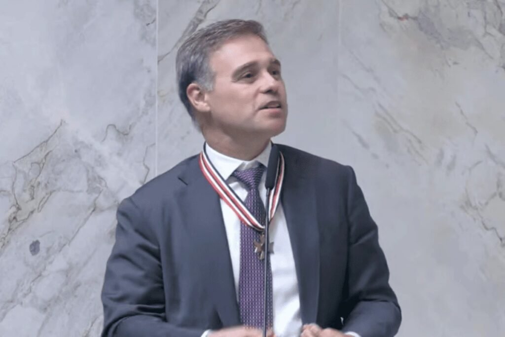 Quando um ministro do **STF** fala em **“não privilegiar amigos e perseguir inimigos”**, a frase não passa despercebida. Por que ela ganhou tanto peso? Porque foi dita por **André Mendonça**, relator dos inquéritos do **caso Master** e das fraudes do **INSS**, em um momento em que sua atuação está no centro de duas investigações de grande repercussão nacional. Onde e em que contexto essa declaração foi feita? A fala ocorreu nesta segunda-feira, **6 de abril**, durante uma cerimônia em homenagem ao ministro na **Assembleia Legislativa de São Paulo**, a **Alesp**. Sem citar diretamente os casos sob sua relatoria, Mendonça afirmou que busca **imparcialidade** e defendeu que não se deve agir para favorecer aliados nem para perseguir adversários. A declaração veio em um discurso de mais de 20 minutos, diante de autoridades de diferentes esferas do poder. Quem estava presente nesse evento? A cerimônia contou com a presença do governador **Tarcísio de Freitas**, do prefeito de São Paulo, **Ricardo Nunes**, e do advogado-geral da União, **Jorge Messias**, indicado pelo presidente **Luiz Inácio Lula da Silva** para uma vaga no Supremo. Também participaram o presidente da Alesp, **André do Prado**, e o presidente do **Tribunal de Justiça de São Paulo**, desembargador **Francisco Eduardo Loureiro**. Mendonça recebeu o **Colar da Honra ao Mérito Legislativo**, homenagem proposta pelo deputado estadual **Oseias de Madureira**. Mas por que as palavras do ministro foram lidas como recados? Porque, além de falar em imparcialidade, Mendonça também abordou a conduta de magistrados. O que ele disse? Afirmou que juízes precisam ter uma **“prudência maior”** em suas relações do que os políticos, sob risco de gerar **“falta de compreensão sobre sua conduta”**. A observação surge em meio a uma crise de **credibilidade** no Judiciário e em um cenário em que ministros do Supremo estão sob questionamento. Quais ministros estão no foco desse ambiente de pressão? **Alexandre de Moraes** e **Dias Toffoli**, colegas de Mendonça no STF, aparecem na berlinda por causa de relações com o banqueiro **Daniel Vorcaro**, ex-dono do **Banco Master**. No caso de Moraes, o Master firmou um contrato de **R$ 129 milhões** com o escritório de advocacia de sua esposa. Esse escritório é suspeito de ter trocado mensagens com Vorcaro no dia em que o banqueiro foi preso pela **Polícia Federal**. Já Toffoli foi sócio de uma empresa que vendeu cotas de um resort no Paraná a um fundo de investimentos ligado a Vorcaro. E por que isso aumenta ainda mais a atenção sobre Mendonça? Porque em Brasília existe a expectativa de que uma eventual **delação premiada** de Vorcaro, cuja homologação cabe a Mendonça no STF, possa atingir os dois ministros. Isso ajuda a explicar por que cada manifestação pública do relator vem sendo observada com tanto cuidado. Como a atuação de Mendonça tem repercutido politicamente? Por ter sido indicado ao Supremo por **Jair Bolsonaro**, suas decisões nos casos do Master e do INSS vêm sendo capitalizadas por bolsonaristas, embora as apurações envolvam personagens de diferentes matizes ideológicos, inclusive ligados ao ex-presidente. As investigações sob sua responsabilidade têm sido usadas por esse grupo para atacar dois de seus principais alvos: **Alexandre de Moraes** e **Lula**. Quais decisões recentes reforçaram esse cenário? A **prisão de Daniel Vorcaro** e a **quebra dos sigilos bancário e fiscal de Lulinha** serviram de munição para o bolsonarismo. Ao mesmo tempo, a presença de autoridades de perfis distintos na homenagem mostrou que a figura de Mendonça hoje ultrapassa um único campo político. E como ele foi tratado durante a sessão solene? Sua trajetória e seu currículo foram exaltados por **Tarcísio de Freitas**, **André do Prado** e por deputados ligados à **Assembleia de Deus Ministério de Madureira**, como **Cezinha de Madureira** e **Oseias de Madureira**. Já **Ricardo Nunes**, que tem feito críticas à postura de ministros do STF em meio ao escândalo do Master, afirmou que a relatoria das investigações está em **“boas mãos”**. O que exatamente disse o prefeito? Nunes declarou que os casos estão com alguém que fará justiça, sem perseguição e sem decisões fora da lei e da Constituição. E foi nesse ambiente, cercado por elogios, pressões e expectativas, que André Mendonça deixou registrada a frase que concentrou as atenções: busca **imparcialidade**, defende **prudência maior** para juízes e sustenta que não se deve **“privilegiar amigos e perseguir inimigos”**.